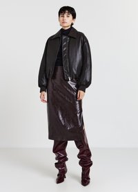 Veste bomber en cuir noir avec fermeture éclair, associée à une jupe midi brillante marron avec une fente sur le côté et des bottes bordeaux montantes.
