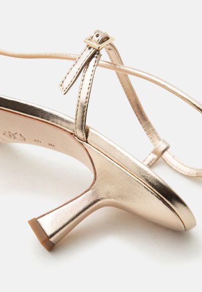 Sandale aurii metalice cu bretele, toc bloc scăzut și închidere cu cataramă la gleznă, prezentate dintr-un unghi lateral pe fundal alb.