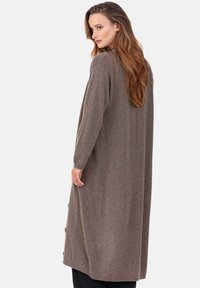 Cardigan long en maille marron, coupe ample, avec un col montant et des boutons latéraux. La texture semble douce et chaude.