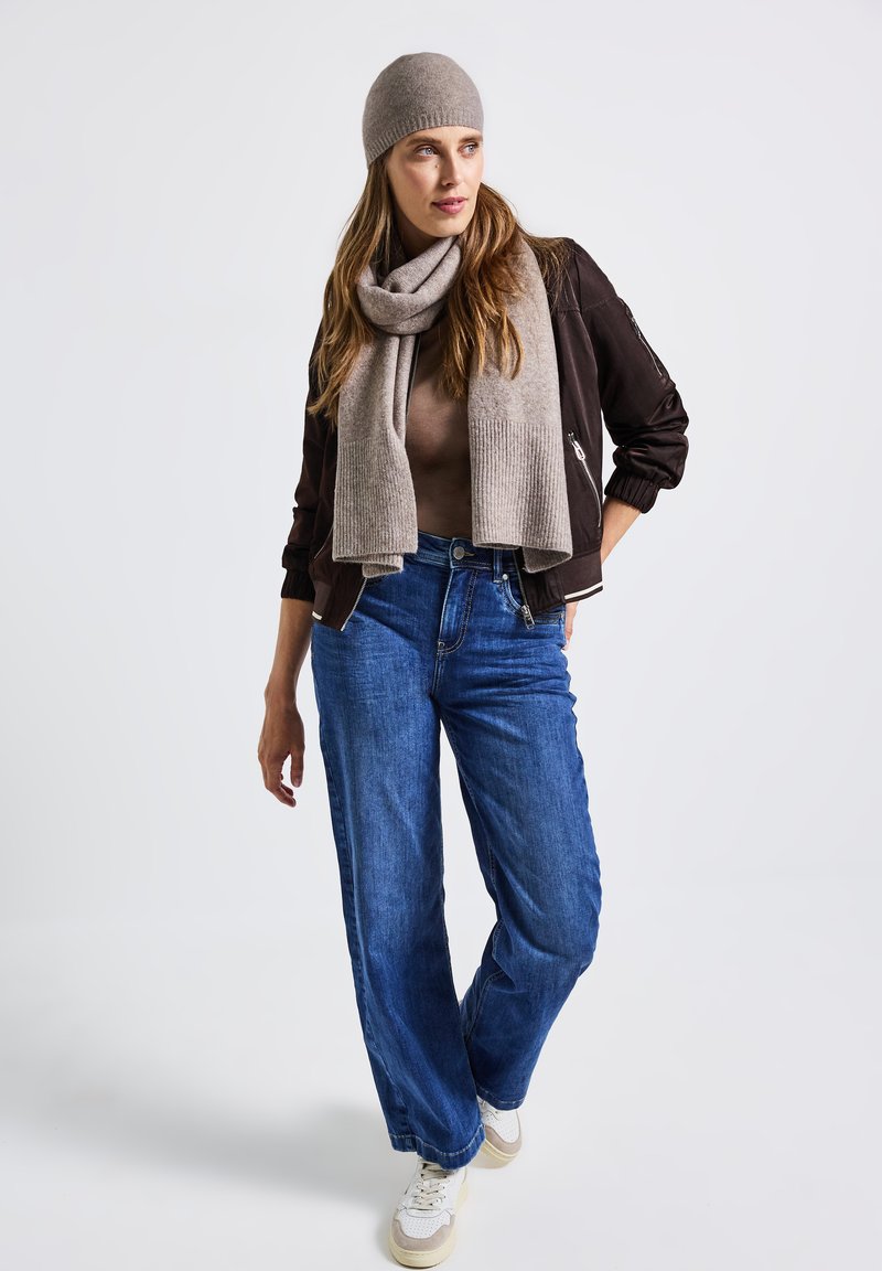 Veste bomber marron, écharpe beige texturée et bonnet assorti. Jean bleu à jambes larges et baskets blanches complètent la tenue.