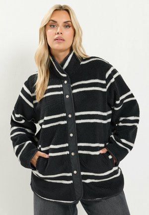 Femme blonde portant une veste en polaire noire avec des rayures horizontales blanches, les mains dans les poches, debout devant un fond clair uni.