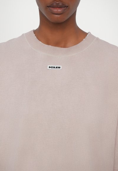 Light pink sweatshirt with a round neck and frayed neckline. Features a small black KSUBI logo tag on the front. Soft texture.