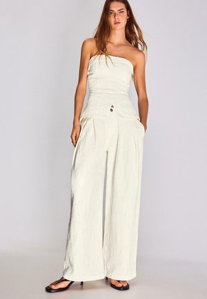 Kobieta ubrana w biały, szerokonaręczny kombinezon bez ramiączek z kieszeniami oraz czarne sandały na obcasie z kwadratowymi noskami, stojąca na jasnym, jednolitym tle.