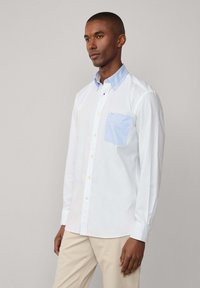 Camicia bianca con colletto azzurro e tasca a quadretti blu. Il tessuto sembra liscio con una vestibilità classica. Pantaloni beige indossati.