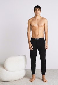 Pantalon noir en coton avec une taille élastique arborant le logo "DIESEL", porté avec un torse nu, debout sur un tapis clair.