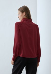 Burgundy blouse with long sleeves, high collar, and buttoned cuffs. Smooth fabric with a fitted silhouette and back seam detailing.