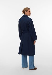 Γυναίκα που στέκεται πλαγιαστά φορώντας ένα σκούρο μπλε παλτό με ζώνη, ανοιχτό μπλε τζιν με πλατιά μπατζάκια και καφέ παπούτσια σε λευκό φόντο.