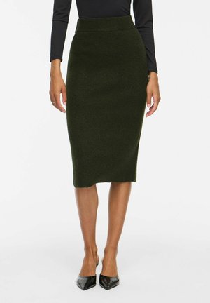 Mujer con falda lápiz de punto verde oscuro, camiseta negra de manga larga y tacones altos negros de punta afilada, de pie frente a un fondo liso.
