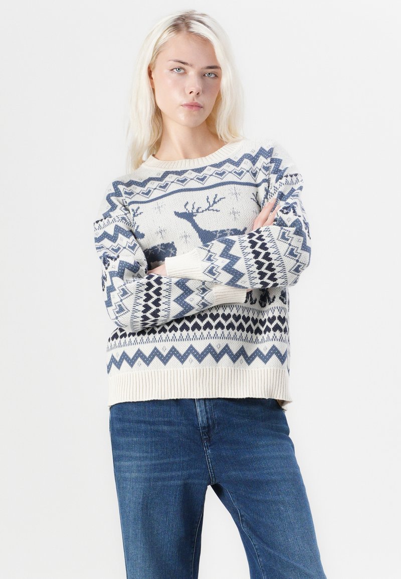 Strickpullover mit einem cremefarbenen Grundton sowie blauen Rentieren und geometrischen Mustern. Gerippte Bündchen und Saum, lockere Passform. Kombiniert mit dunkelblauen Jeans.