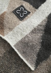 Wollen deken in grijstinten, beige en wit met een decoratief stoffen label met een bloemenembleem en een zachte, textuurige oppervlak.