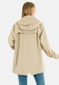 Chaqueta impermeable de color beige claro con capucha, corte holgado, costuras visibles y un dobladillo ligeramente curvado. Textura suave y diseño minimalista.
