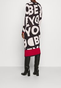 Robe en maille ornée de grandes lettres noires et blanches sur un fond sombre, ourlet rouge et manches longues, associée à des bottes noires à talons.