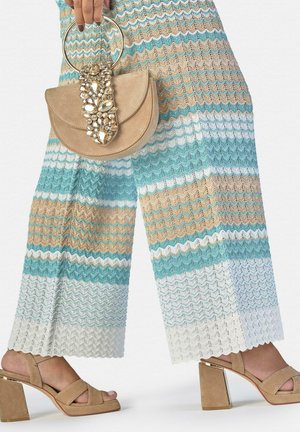 Mano sostiene un bolso de ante beige con detalle dorado con pedrería, vistiendo pantalones de punto a rayas en azul, beige y blanco, y sandalias con tacón bloque beige.