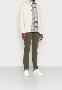 Giacca button-up beige chiaro, camicia a quadri verde e blu sotto, pantaloni verde oliva e sneakers bianchi. Outfit semplice e casual.
