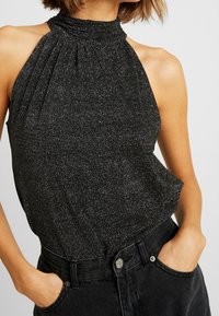 Haut noir à bretelles avec un col froncé, présentant une texture scintillante. Associé à un pantalon en denim noir taille haute, mains dans les poches.