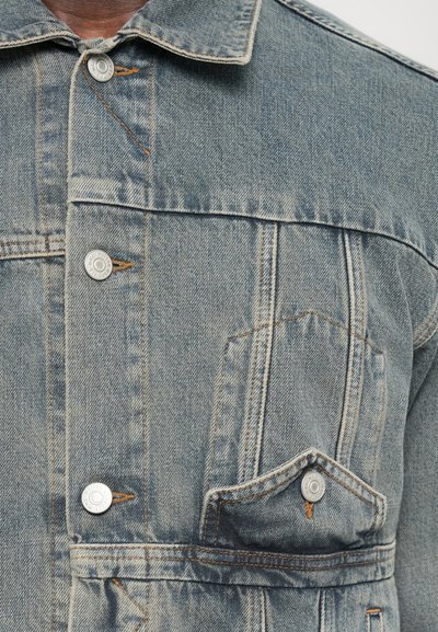 Jachetă din denim de culoare bleu deschis cu nasturi argintii, guler ascuțit, buzunar pe piept și cusături portocalii în contrast. Textura este uzată și ușor decolorată.