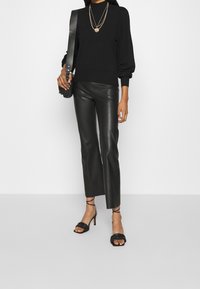 Schwarzer Pullover mit Puffärmel, kombiniert mit schwarzen Lederhosen und schwarzen Sandaletten mit Riemchen. Dreischichtige goldene Halsketten sorgen für zusätzliche Details.