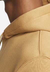 Světle hnědá mikina s texturovanou tkaninou, která má kapuci na šňůrku a švy podél ramen. Detailní záběr na oblast výstřihu.