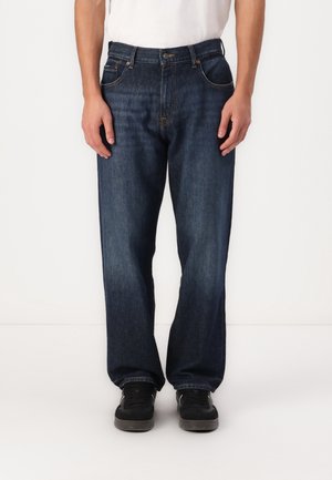 Personne portant un jean droit bleu foncé et des baskets noires, debout devant un fond blanc uni.