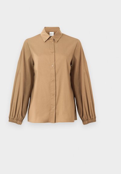 Blouse beige à manches longues avec boutonnière sur le devant, col pointu, poignets élastiques froncés et coupe légèrement ample.