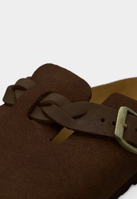 Καφέ σουέτ σανδάλι με πλεκτό λουράκι και μεταλλική αγκράφα. Στρογγυλεμένο σχήμα με ανοιχτές πλευρές και υφασμένη επιφάνεια.