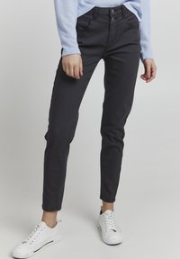 Zwarte slim-fit jeans met een dubbele knoopsluiting, voorzakken en een gladde textuur, gecombineerd met witte sneakers en een lichtblauw shirt.
