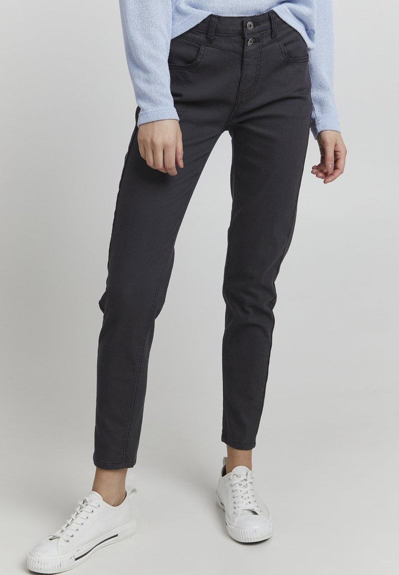 Zwarte slim-fit jeans met een dubbele knoopsluiting, voorzakken en een gladde textuur, gecombineerd met witte sneakers en een lichtblauw shirt.