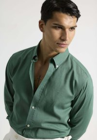 Camisa verde de botón en tejido suave, con mangas largas y cuello. Los detalles notables incluyen cierres de botón y un logo sutil.