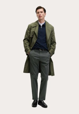 Man draagt een groene trenchcoat, marineblauwe trui met quarter-zip, grijze broek, zwarte riem en zwarte nette schoenen, met handen in de zakken.