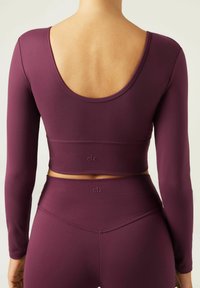 Langärmliges Sport-Crop-Top aus tiefburgunderfarbigem Stoff, mit einem Rundrücken und Logostickerei am Saum, kombiniert mit passenden hochgeschnittenen Leggings.