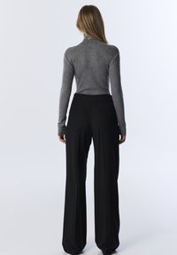 Grau gestrickter Rollkragenpullover mit Verzierungen, kombiniert mit hochtaillierten, weit geschnittenen schwarzen Hosen. Beide Kleidungsstücke zeigen klare Linien.