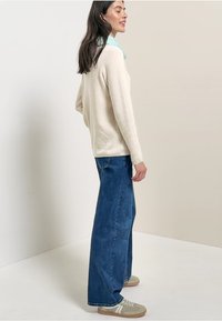 Beige Pullover mit strukturierter Oberfläche, kombiniert mit weit geschnittenen blauen Jeans. Ein hellgrüner Schal sorgt für eine zusätzliche Schicht. Bequeme Sneakers runden den Look ab.