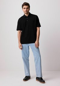 Sort kortærmet polo shirt med lomme, båret med lyseblå jeans og sorte sko. Enkelt design, bomuldsstof, klassisk pasform.