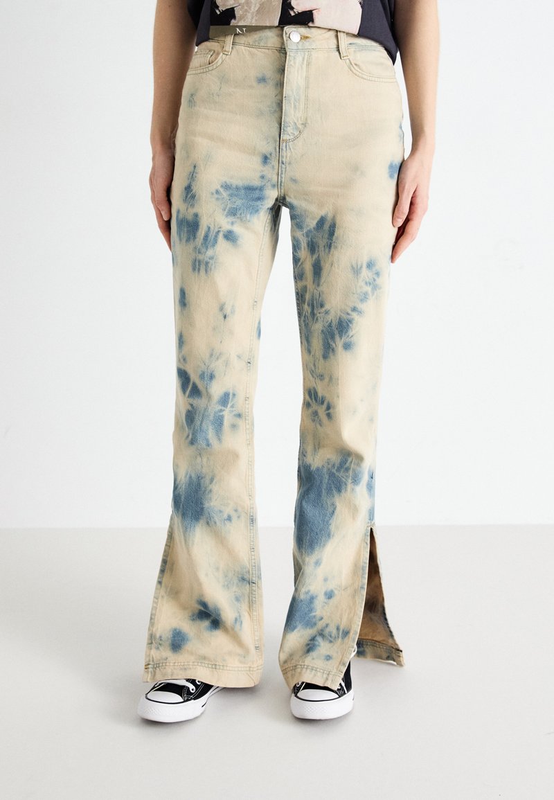 Jeans de color beige deslavado con patrones de tinte anudado azul, piernas acampanadas y aberturas laterales. Llevados con zapatillas de deporte de patrón en blanco y negro.