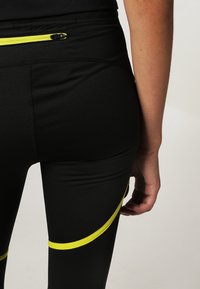 Černé legíny vyrobené z pružného materiálu, se úzkou žlutou zipovou kapsou na zadní straně a žlutým akcentem podél stehna.