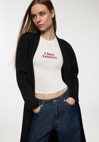 Cardigan nero lungo su una maglietta crop bianca con "Ciao Amore" in rosso, abbinato a jeans di denim blu scuro. Design semplice e aderente.