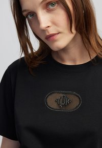 Camiseta negra con un parche ovalado de malla y acentos brillantes. Mangas cortas y cuello redondeado clásico. Textura de tela suave.