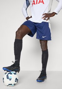 Maglia da calcio Nike con una maglietta bianca con scritte rosse, pantaloni corti blu navy, calze nere e scarpini neri su un pallone da calcio con un motivo.