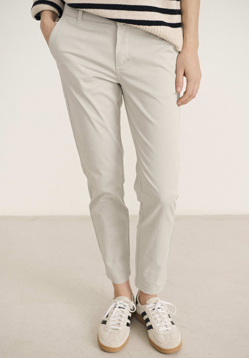 Pantalon chino beige clair avec une coupe slim et des poches avant, associé à des baskets blanches et beiges avec des rayures noires.