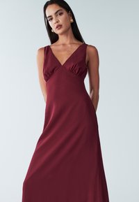 Vínové šaty bez rukávov s hlbokým výstrihom do V, s nariaseným detailom na prsiach, plynúcou látkou a hladkou textúrou.