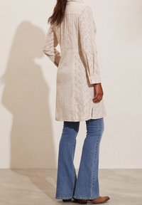 Tunic lunga color beige chiaro a maniche lunghe con motivo a occhio di pernice; abbinata a jeans blu a campana e stivaletto brown; presenta un colletto e un'apertura laterale.