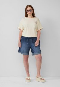 T-shirt crème à manches courtes avec un design de soleil, associé à un short en denim foncé et des sandales à plateforme de couleur claire. Le mannequin porte des lunettes de soleil.