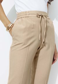 Pantalons beiges à cordon de serrage en tissu lisse avec une taille élastique. Comprend deux poches latérales et un design de jambe fuselée.