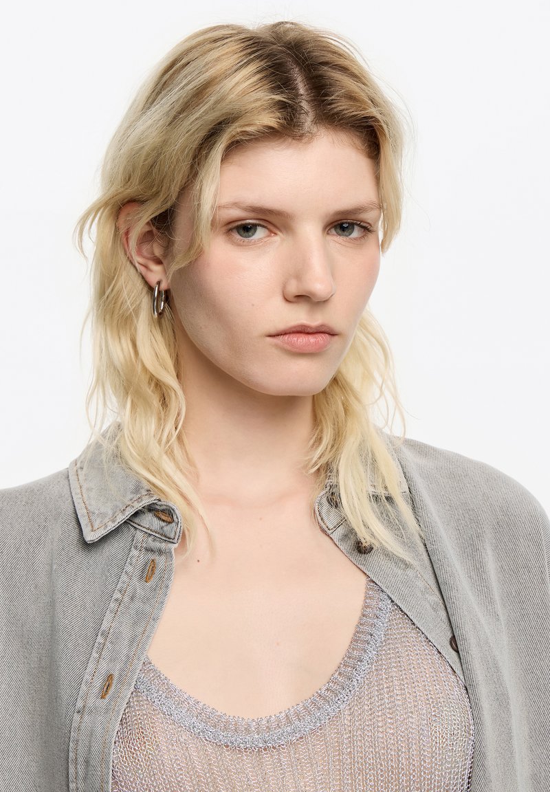 Szara koszula z denimu z guzikami na warstwie cienkiego topu ze srebrną nitką. Modelka ma falowane, sięgające ramion blond włosy. Minimalne dodatki.