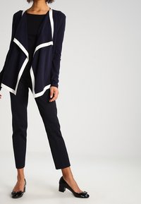 Cardigan blu navy con rifiniture bianche, frontale drappeggiato, top aderente nero, pantaloni neri testurizzati e tacchi in vernice nera, in posa in piedi.