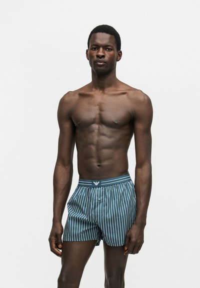 Άνδρας χωρίς πουκάμισο στέκεται φορώντας μπλε και άσπρο κάθετα ριγέ μπόξερ με λογότυπο στη μέση, μπροστά σε απλό ανοιχτό φόντο.