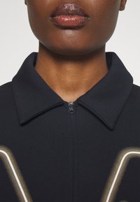 Giacchino zip-up di un blu navy scuro con colletto, texture a coste e una grafica bianca e dorata sul davanti.