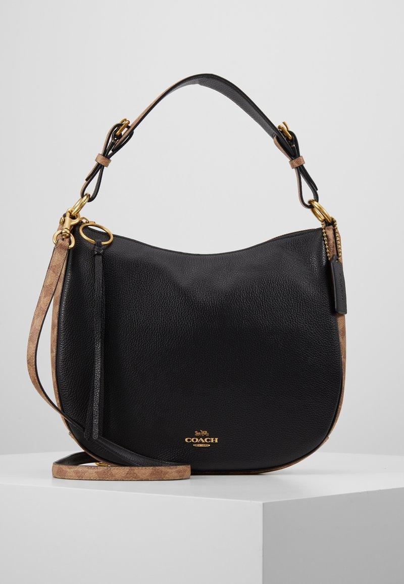 Sac à main Coach en cuir noir avec forme incurvée, ferrures dorées, accents marron, poignée courte et longue bandoulière réglable sur surface blanche.
