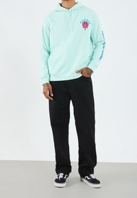 Mintgrön hoodie med en framficka, rosa grafik och text på bröstet, samt blå text på ärmen. Matchad med svarta byxor och sneakers.