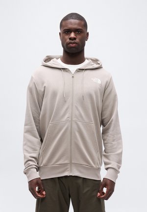 Jonge man met een beige North Face hoodie met rits en donkere broek, staand tegen een effen lichte achtergrond, naar voren gericht.